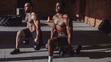 2 durchtrainierte Männer machen im Gym mit Kettlebells in den Händen Ausfallschritte.