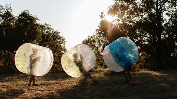 Drei Personen stecken in großen, transparenten Bällen und laufen über eine Wiese im Sonnenuntergang.