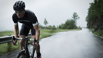 Ein Mann in Radsport-Kleidung fährt im Stand auf einem Rennrad. Hinter ihm erstreckt sich eine nasse Straße.