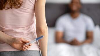 Ein Mann sitzt im Bett, davor steht seine Freundin und versteckt einen positiven Schwangerschaftstest hinter ihrem Rücken