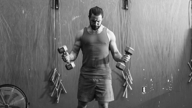 Ein Mann steht in einem Gym und hebt zwei Kurzhanteln. Er blickt konzentriert zu Boden.