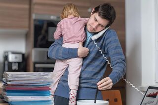 Ein Vater beim Multitasking in der Küche mit seinem Kind auf dem Arm und einem Telefonhörer unters Kinn geklemmt