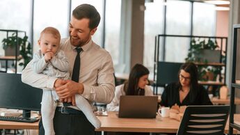 Ein Vater im Business-Outfit hält in einem Büro ein kleines Kind auf dem Arm, im Hintergrund sieht man Kolleginnen bei der Arbeit