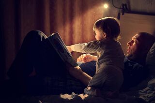 Ein Vater liegt mit seinem Kind im Bett und liest ihm aus einem Buch vor