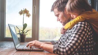 Ein Vater sitzt an seinem Laptop, sein Sohn schaut ihm über die Schulter