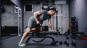 Ein muskulöser Mann trainiert mit einer Hantel, die sich auf einer Sportbank im Fitnessraum befindet. 