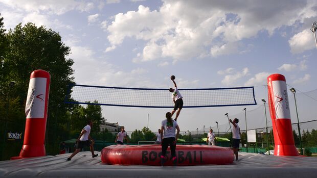 Eine Gruppe Männer und Frauen befinden sich auf einem aufblasbaren Spielfeld mit einem Netz in der Mitte. Eine Person springt und versucht, an den Ball zu gelangen.