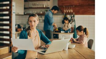 Eine Mutter sitzt am Esstisch in der Küche und arbeitet am Notebook, im Hintergrund sieht man die zwei Töchter und den Mann, der gerade das Essen zubereitet