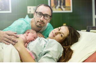 Eine Wöchnerin liegt mit ihrem Baby auf dem Bauch in einem Krankenhausbett, der Vater des Kindes sitzt daneben und umarmt die beiden