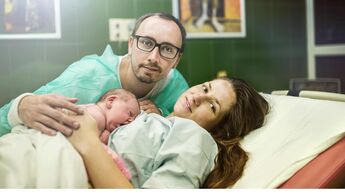 Eine Wöchnerin liegt mit ihrem Baby auf dem Bauch in einem Krankenhausbett, der Vater des Kindes sitzt daneben und umarmt die beiden