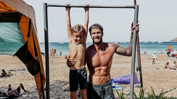 Matty Fox und sein Sohn an einem Strand. Der Kleine hängt an einer Klimmzugstange, während Matt ihn von der Seite hält. 