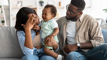 Mutter und Vater sitzen auf einem Sofa. Die Mutter hat ein schreiendes Kleinkind auf dem Arm, das sich vom Vater abwendet