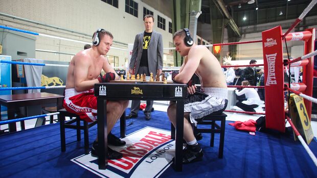 Zwei Männer in Boxshorts sitzen in einem Boxring an einem Schachbrett.