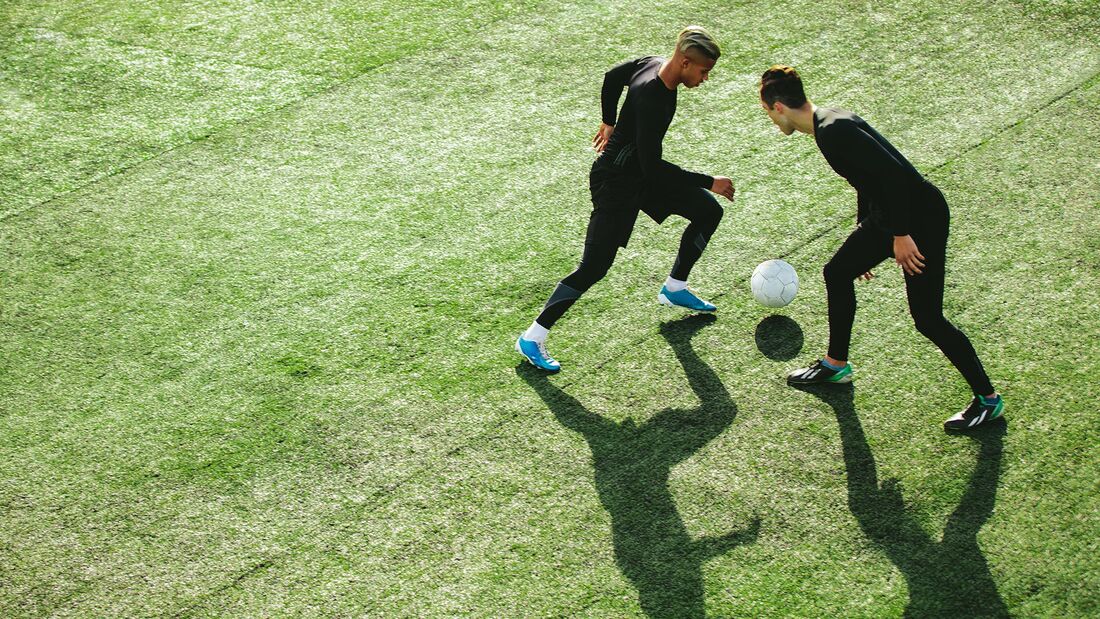 Zwei Männer in dunklen Outfits spielen gegeneinander Fußball