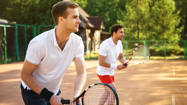 Zwei Männer mit weißen T-Shirts stehen an der Grundlinie und halten jeweils einen Tennisschläger in der Hand.